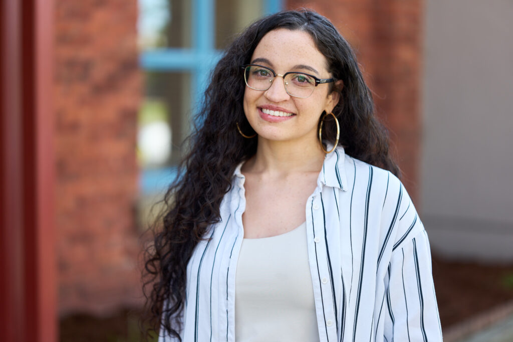 Samantha is pictured in front of a blurry brick background, smiling in a white striped button down shirt, glasses, and gold hoops