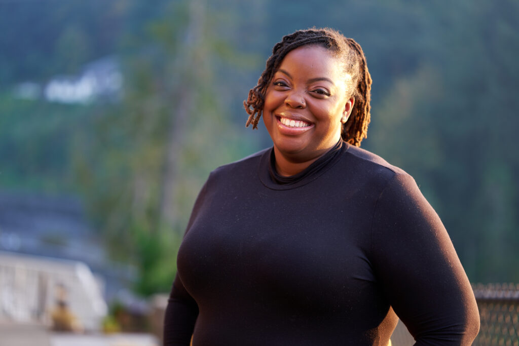 Maya, wearing a black shirt and smiling in front of a blurred tree-filled background.