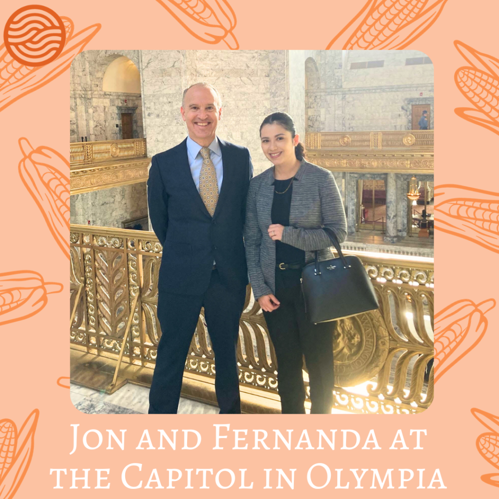 Jon and Fernanda stand smiling, side by side, in front of a golden colored handrail. Jon is dressed in a navy blue suit with a patterned gold tie. Fernanda is wearing a gray blazer, black slacks, a black blouse, and a black purse. Her hair is pulled back in a ponytail. 
The photo is surrounded by a light orange border with darker orange corn husks. In the top right corner is the Pathwaves logo, which resembles to waves coming together like joined palms. 
Below the photo reads "Jon and Fernanda at the Capitol in Olympia" 