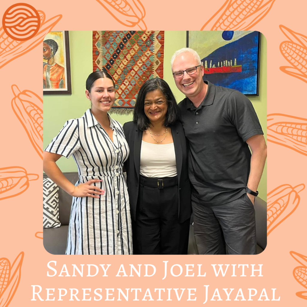 Sandy, Rep. Jayapal, and Joel stand together, smiling with their arms around each other. They stand in front of some abstract paintings which are obstructed from view. The photo is surrounded by a light orange border with darker orange corn husks. In the top right corner is the Pathwaves logo, which is a circle with two connecting waves - resembling hands resting in each other. Below the photo, in white script, reads: "Sandy and Joel with Representative Jayapal". 