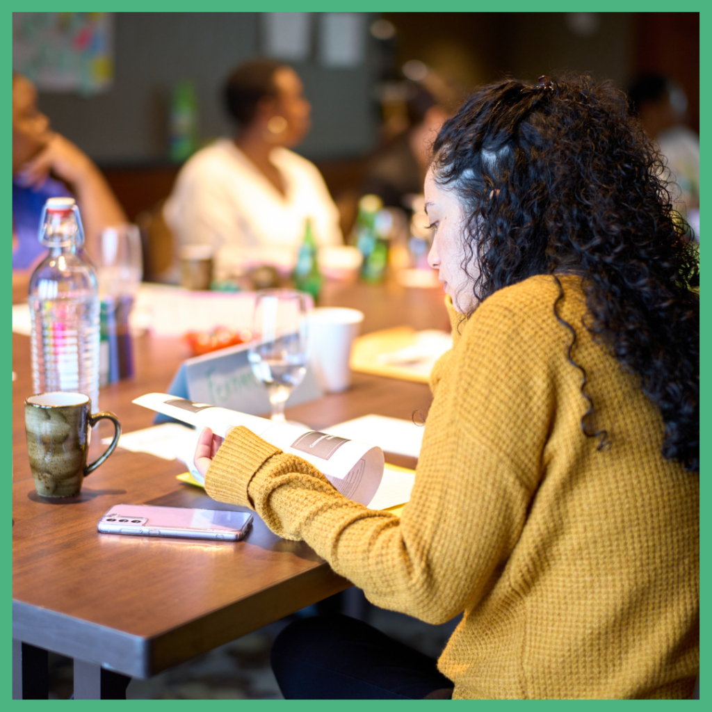 Pathwaves Fellow, Fernanda, sits reading a handout. She is wearing a yellow sweater and has her dark curly hair pulled up in a half ponytail. In the background, you can see other Fellows looking at a speaker who is outside of the frame. 