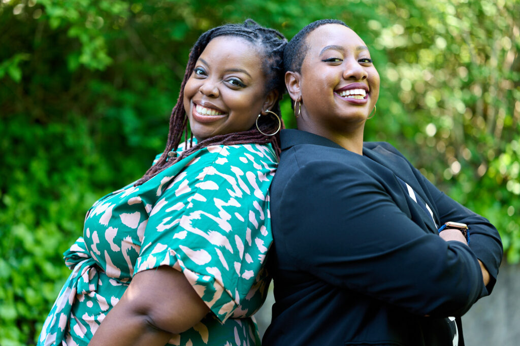 Two Black women standing back to back and smiling looking at the camera in front of a leafy green background.