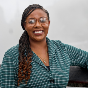 Emani is smiling before a foggy background and wearing glasses, a turquoise blazer, and her hair braided