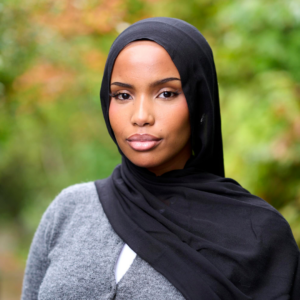 Asha wears a black hijab and gray cardigan, looking at the camera with a blurry green background