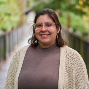 Dania has short brown hair and wears glasses, an earth-tone shirt and cream cardigan, smiling and standing before a wooden bridge and trees