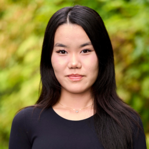 Misheel has black hair and a slight smile on her face, wearing a black top and standing before a leafy green background