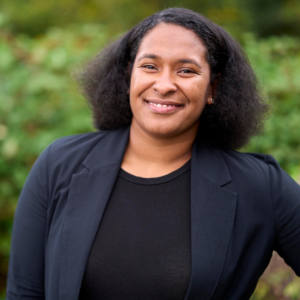 Raishawn smiles in a black top and black blazer in front of a blurry green background