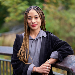 Gemma is wearing a gray button down, black cardigan, and long braids, leaning against a wooden railing with trees in the background