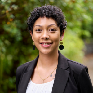 Jasmine is wearing a white top and black blazer with dangly earrings, smiling in front of a green background