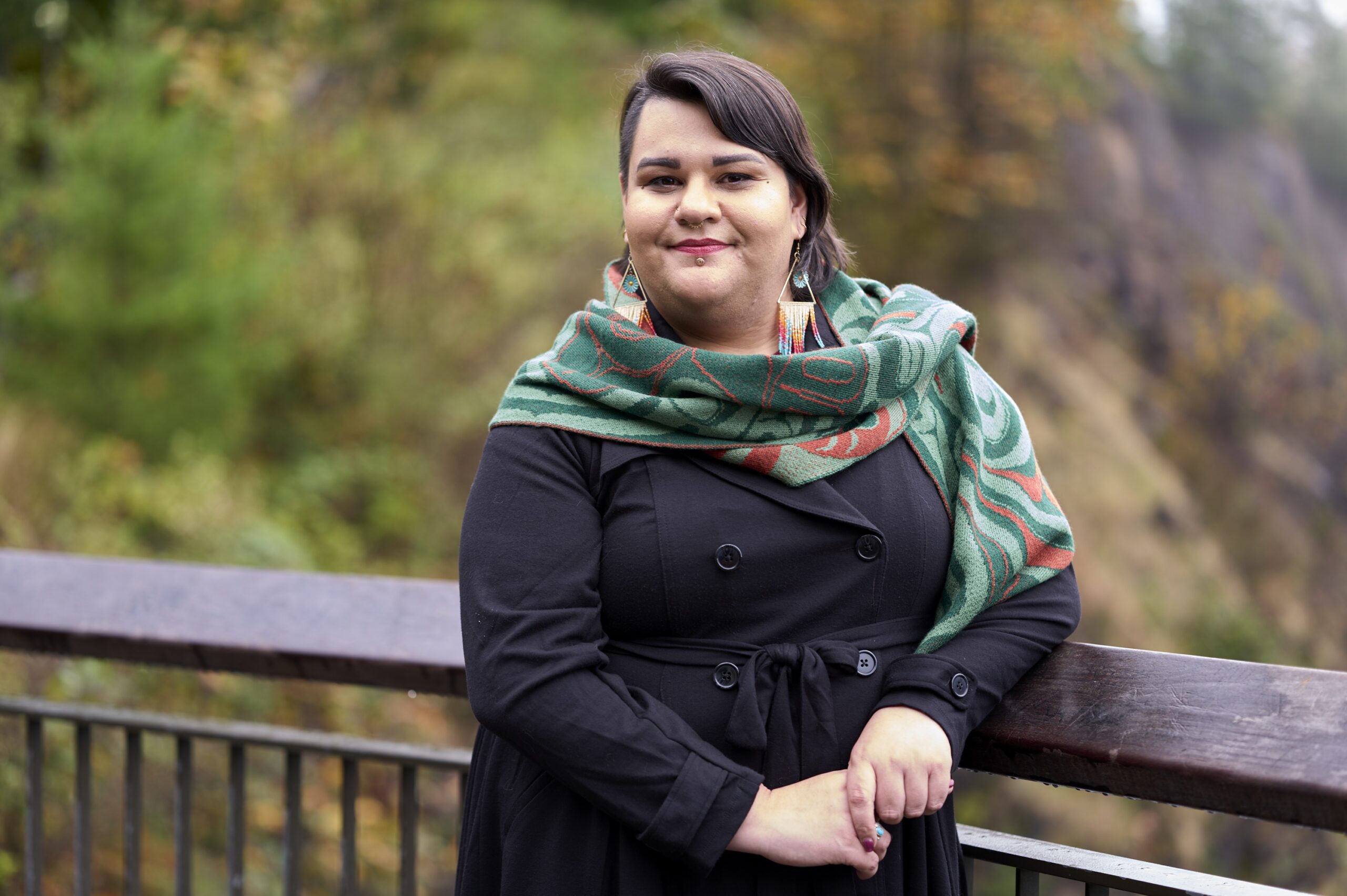 Barbara stands leaning against a wooden railing with rock and greenery behind them. She has brown hair, a stud below her bottom lip, and a green and orange scarf with indigenous designs covering her black trench coat. Barbara's expression is gentle and serious.