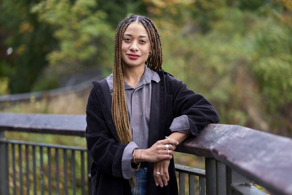 Gemma stands looking at the camere with a serious smile, long braids tucked behind and in front of her shoulder as she rests on a wooden railing. She is wearing a gray top with a black jacket, silver rings and earrings.