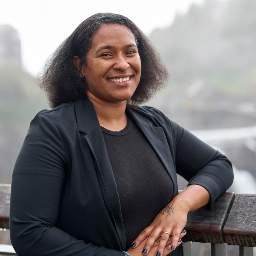 Raishawn stands smiling in front of a gray background. She is wearing a black blazer and black top.
