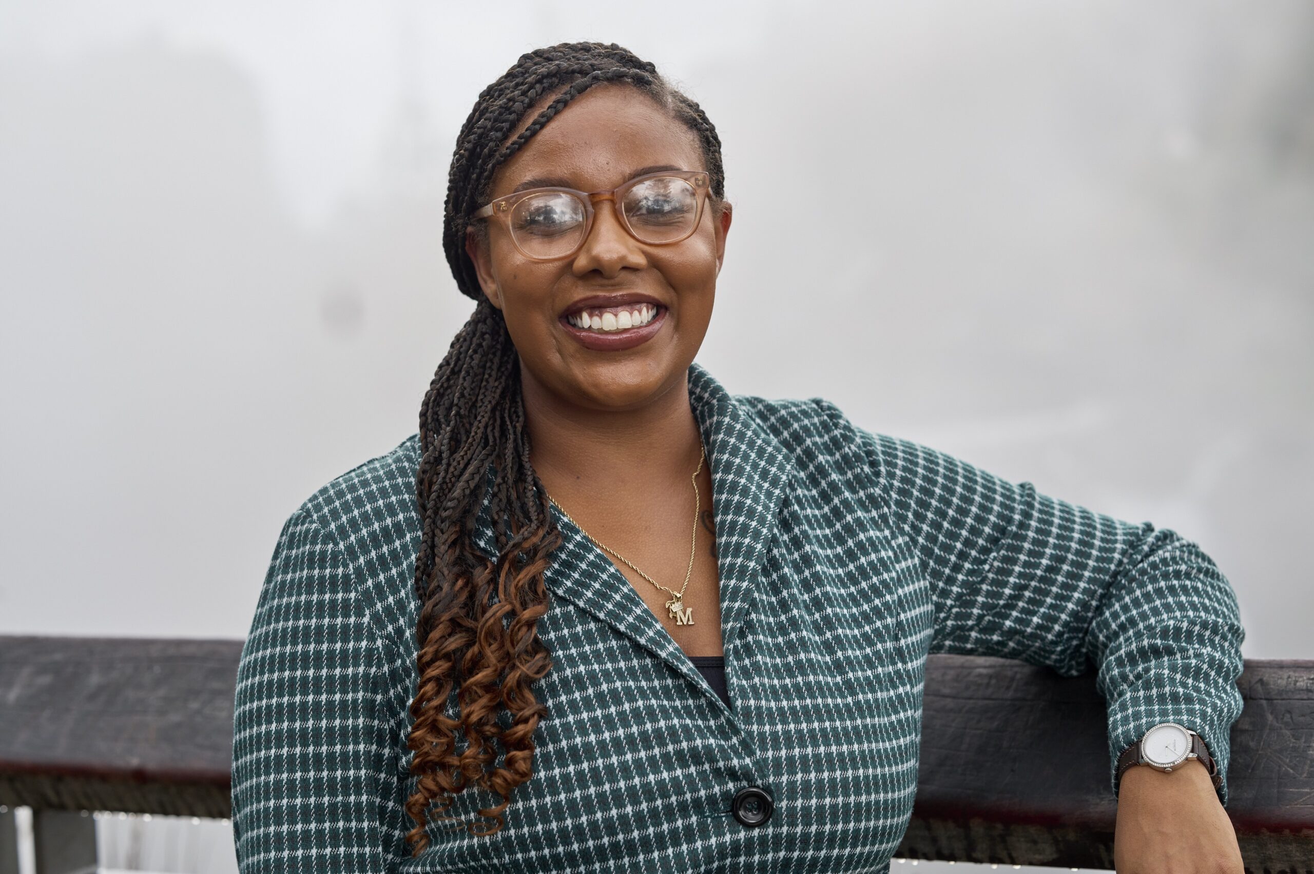 Emani smiles in front of a foggy background. She has long braided hair, glasses, and a patterned teal blazer