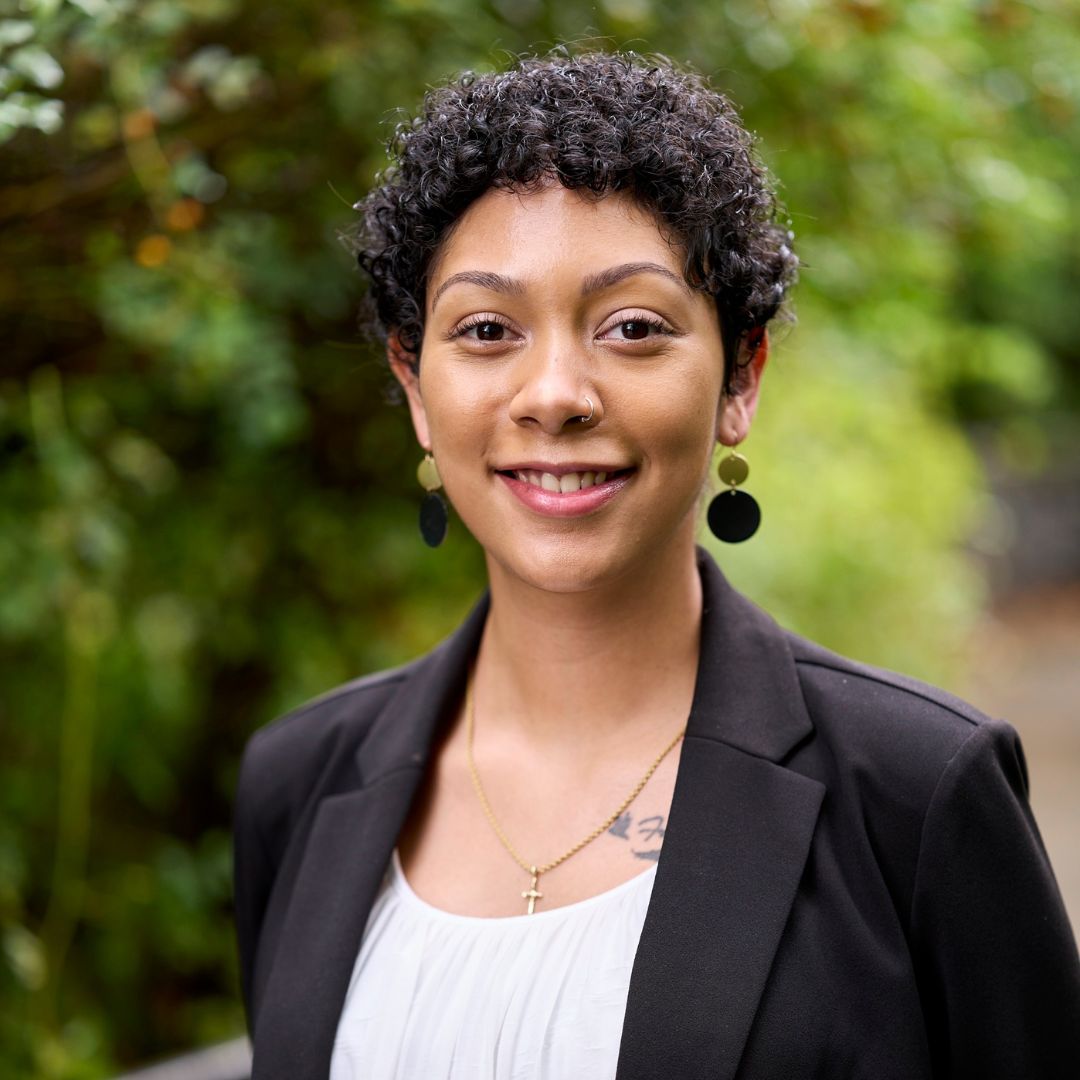 Jasmine smiles in front of a verdant background. Her hair is short and curly and she is wearing a white top with a black blazer, gold cross necklace, and dangle earrings.