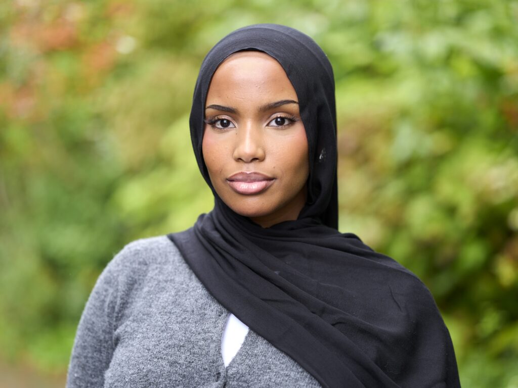 Asha stands in front of a verdant background. She is wearing a black hijab and gray cardigan sweater.