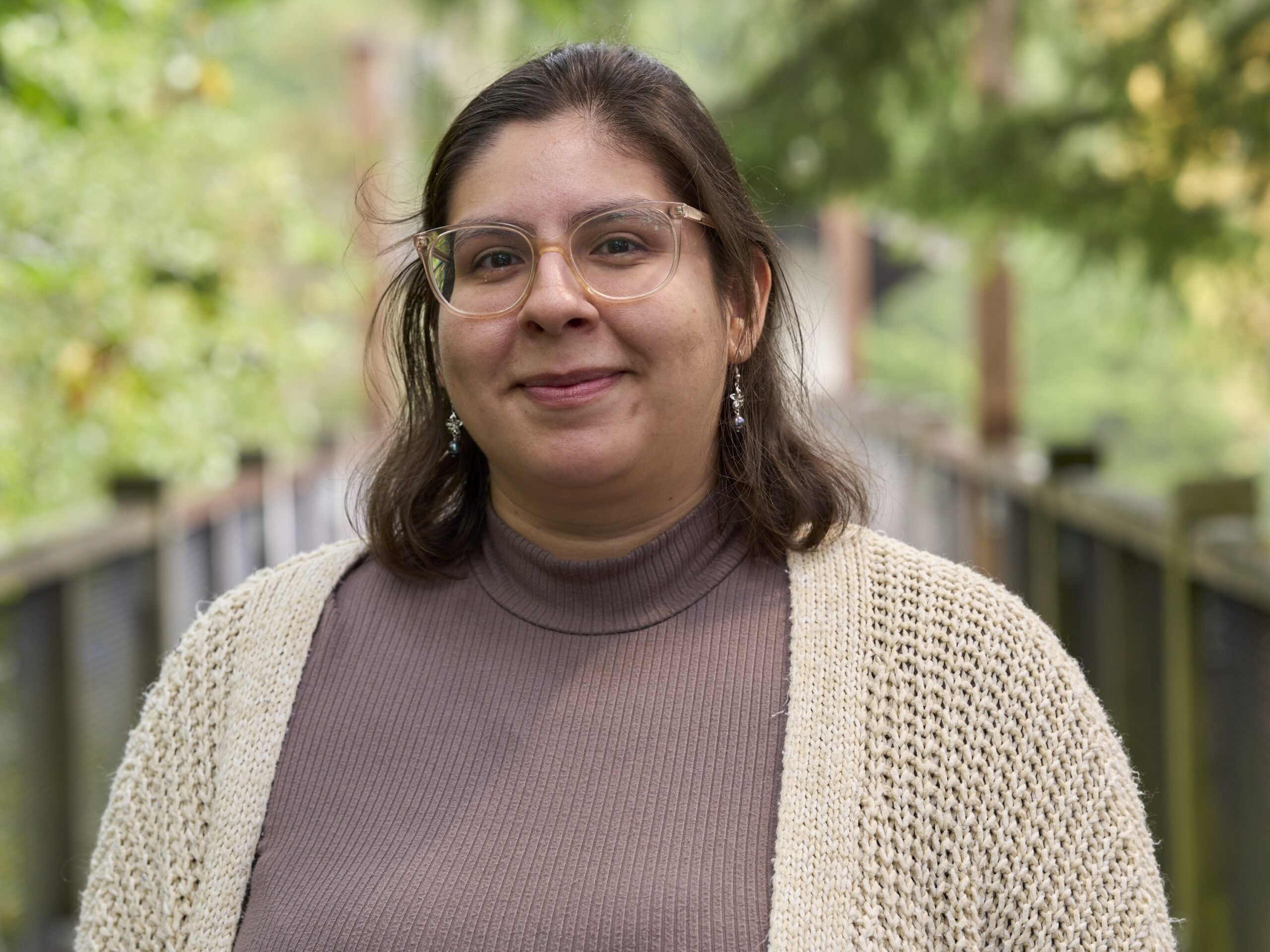 Dania smiles in front of a verdant background. They are wearing glasses and a cream sweater with a mauve turtleneck